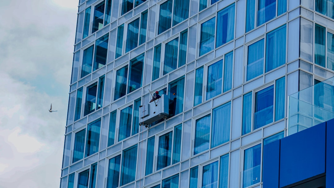 A close-up of a high-rise building in Tel Aviv, featuring a glass facade with modern architectural design. A worker in a suspended platform is cleaning the windows, illustrating urban maintenance and safety equipment. A bird is visible flying in the background, adding a dynamic element to the image. The clear reflection on the glass windows enhances the modern cityscape ambiance.